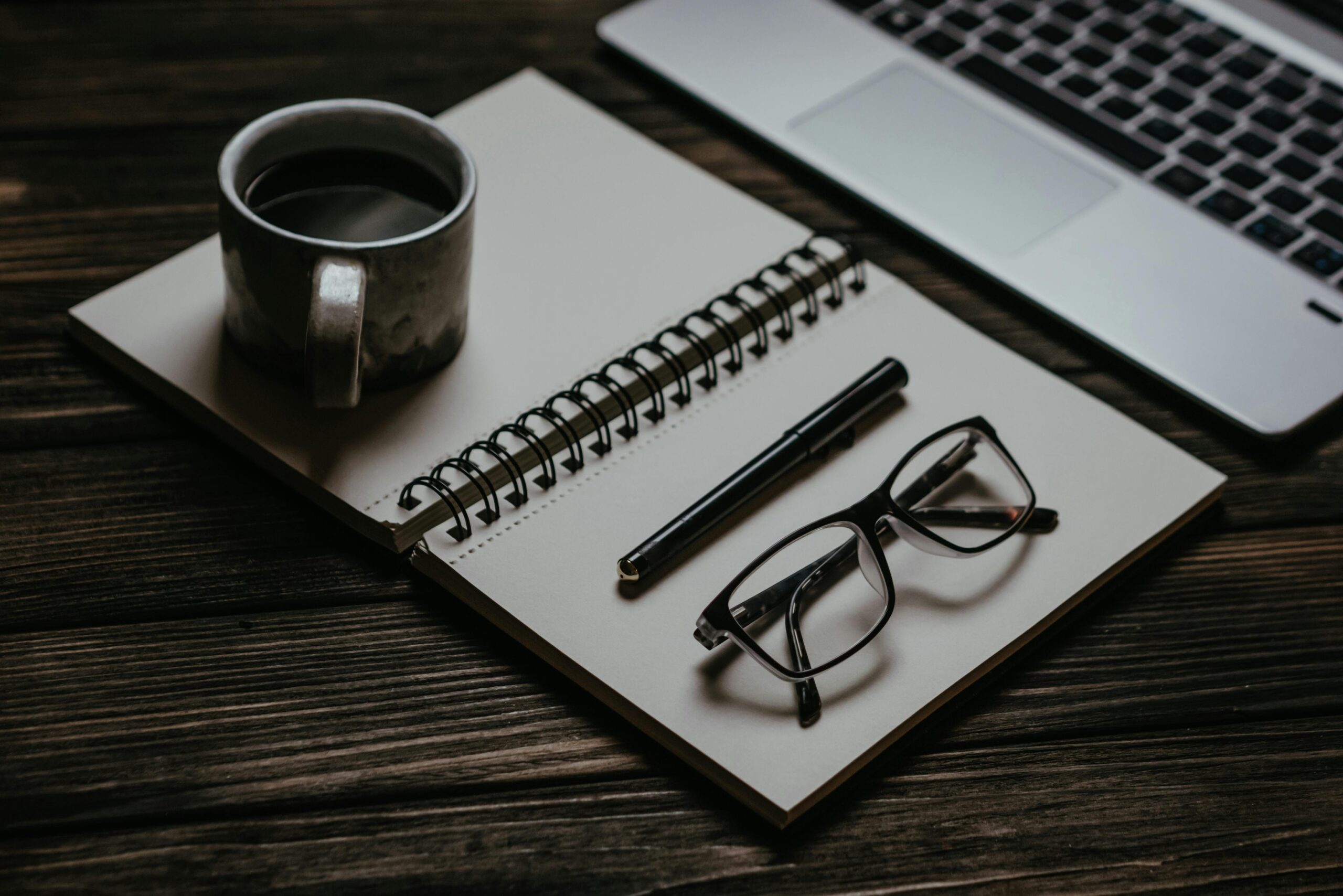 Cozy workspace with a notebook, coffee, glasses, and laptop on a wooden table.
